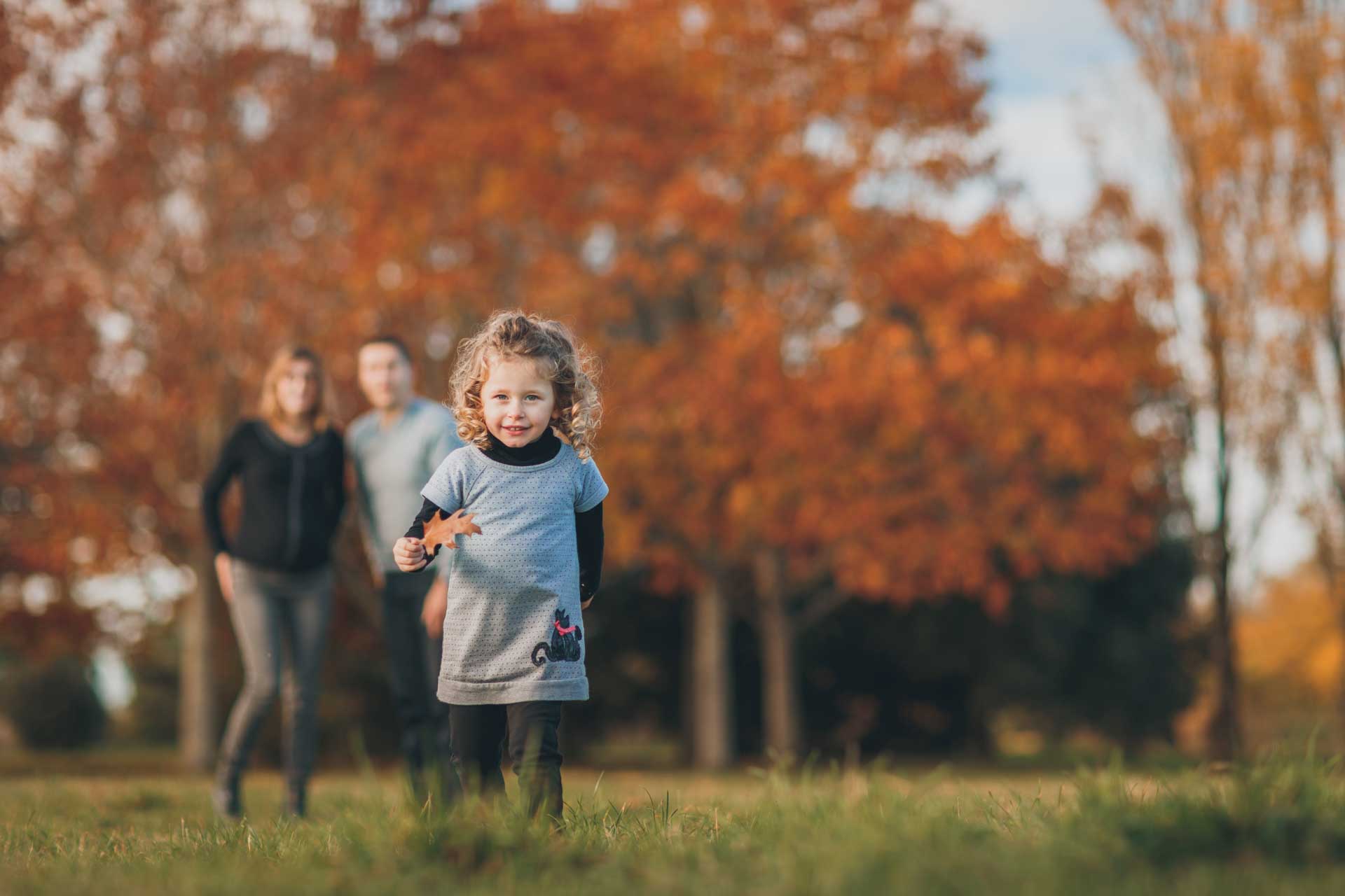 Enfant qui court avec une feuille à la main sous le regard de ses parents en arrière plan, devant des arbres
