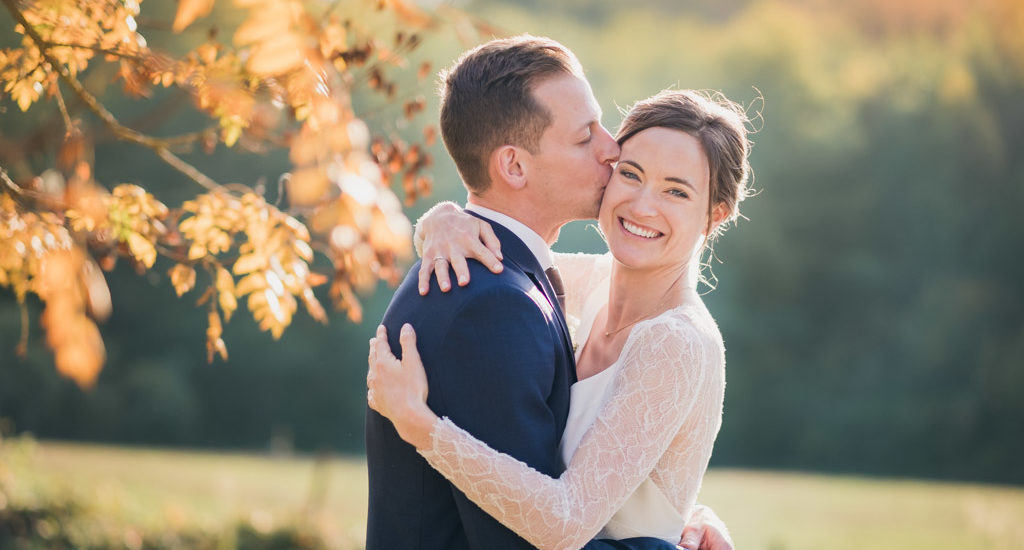 Le marié fait un bisou sur la joue de sa femme souriante et radieuse au milieu des arbres avec des feuilles jaunes d'automne