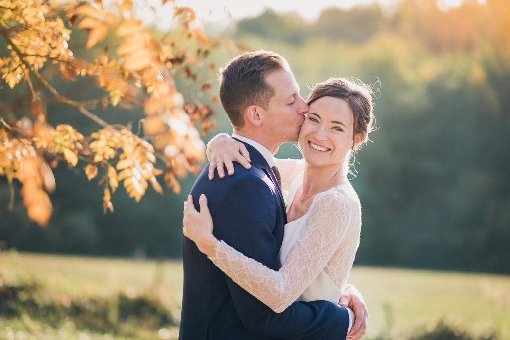 Le marié fait un bisou sur la joue de sa femme souriante et radieuse au milieu des arbres avec des feuilles jaunes d'automne