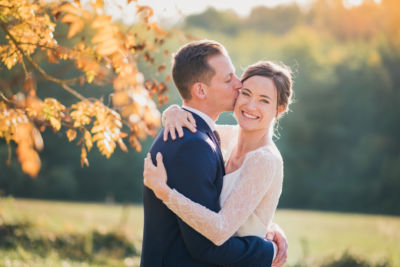Mariée radieuse embrassée par son mari dans un cadre champetre avec de jolies couleurs d'automne dans les arbres