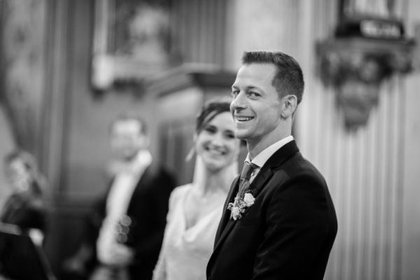 Portrait du marié souriant, on aperçoit sa femme qui le regarde en arrière plan