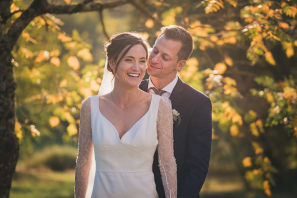 Le marié se tient derrière la mariée et lui chuchote quelque chose à l'oreille qui la fait rire. Ils sont sous des arbres avec des feuilles d'automnes jaunes et oranges.