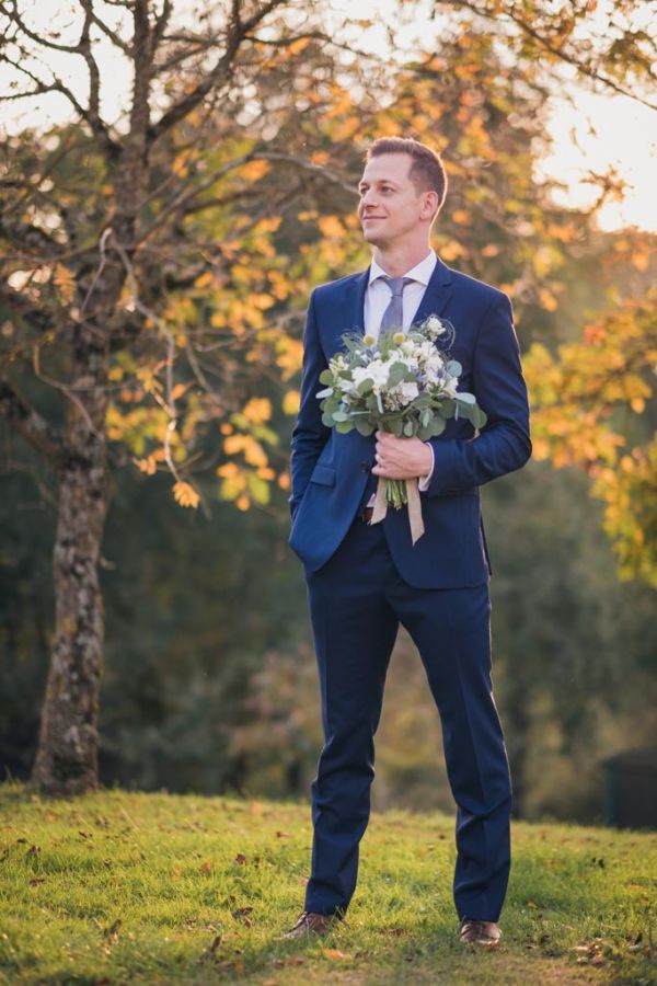 Le marié est debout devant des arbres tenant le bouquet de la mariée dans sa main